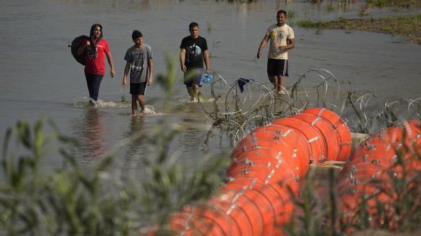 Migrants walk past the site wher<em></em>e workers are assembling large buoys to be used as a border barrier along the banks of the Rio Grande in Eagle Pass, Texas, Tuesday, July 11, 2023.
